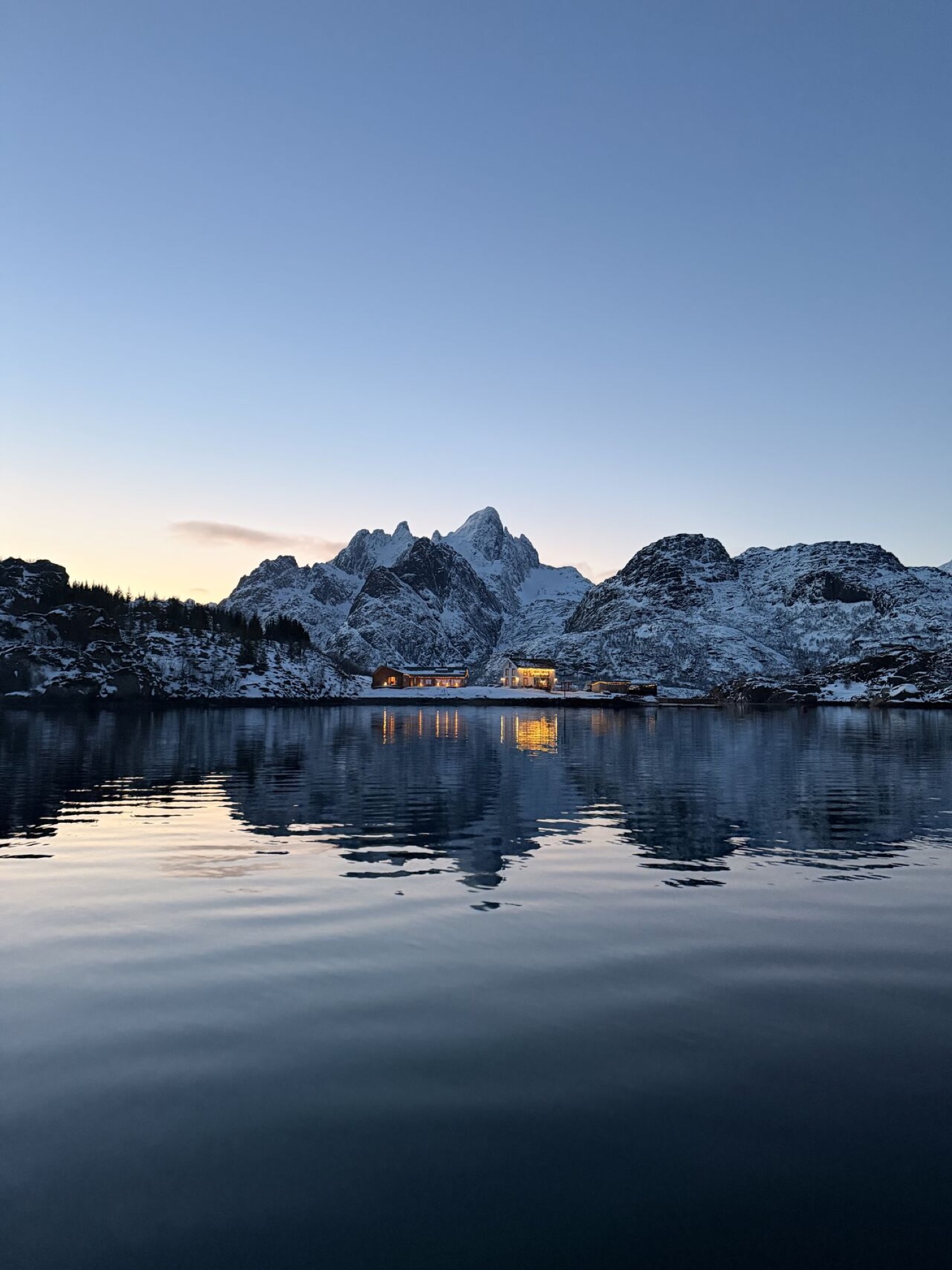 Brakøya i Lofoten i blått kveldslys, med opplyste bygninger som speiler seg i fjorden under snøkledde fjell