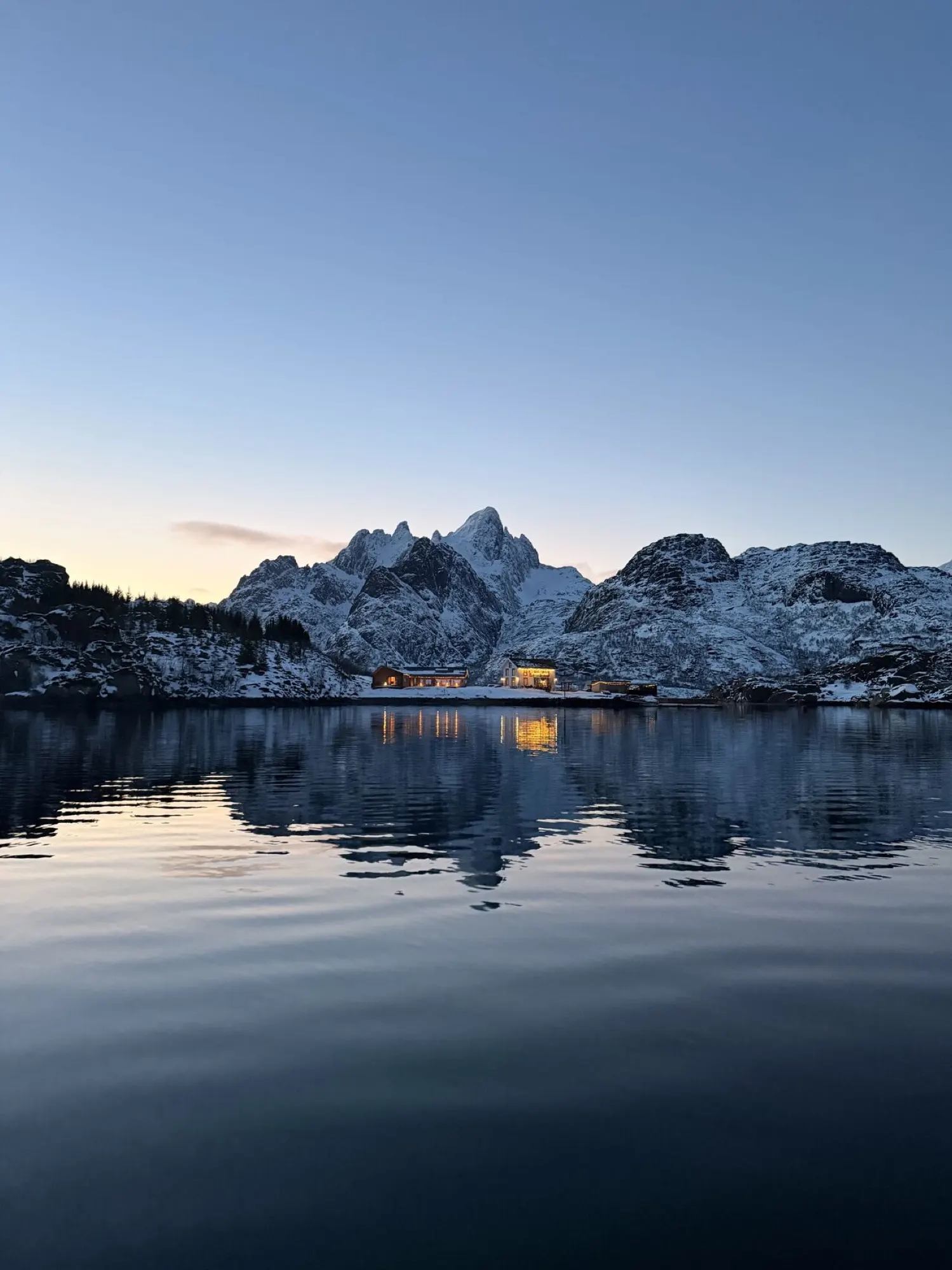 Brakøya i Lofoten i blått kveldslys, med opplyste bygninger som speiler seg i fjorden under snøkledde fjell