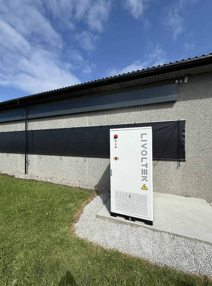 Hvitt batteriskap fra Livoltek på et betongfundament langs veggen på en driftsbygning, med gress og blå himmel rundt