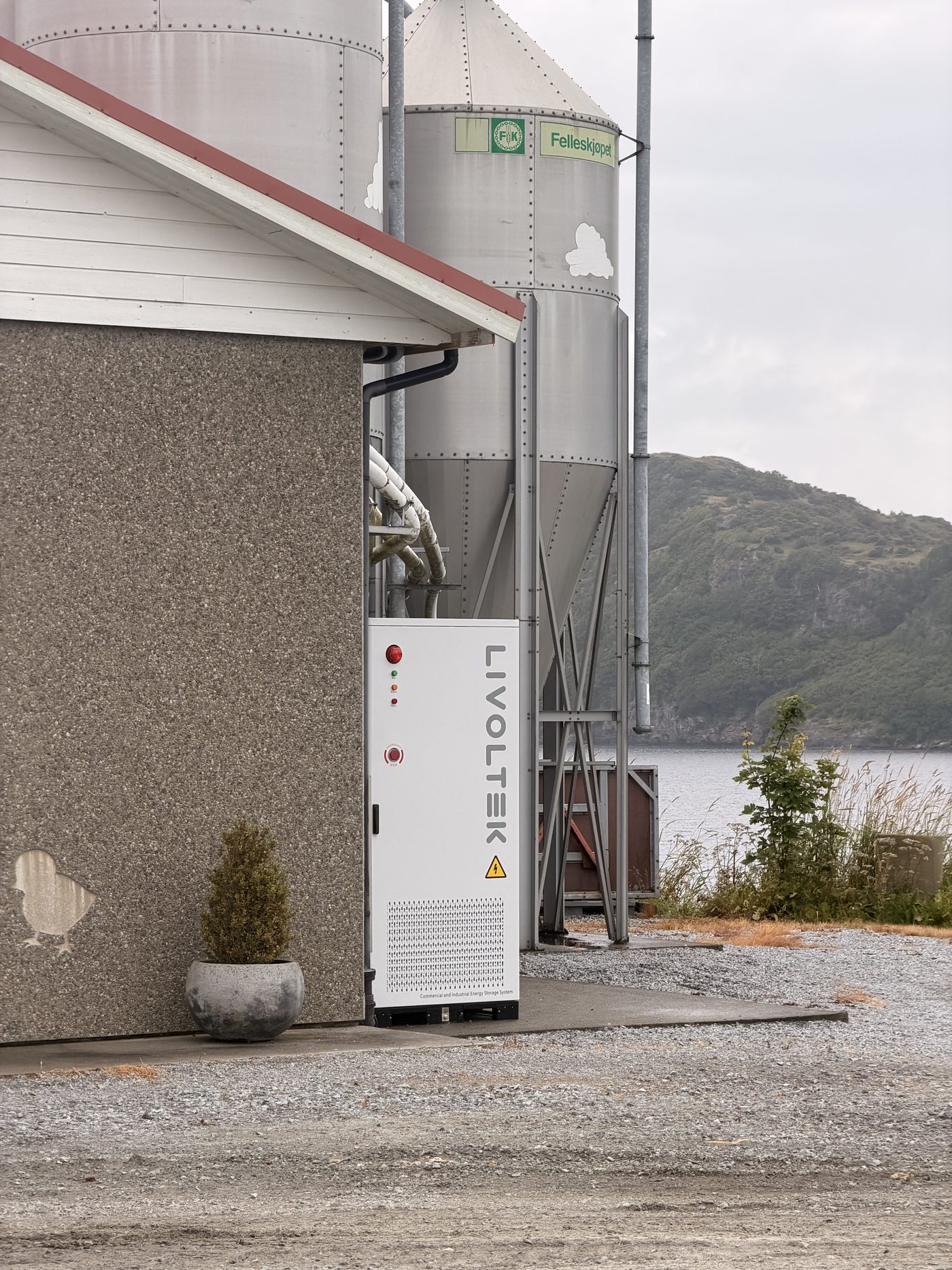 Det hvite batteriskapet ved veggen på kyllinghuset, med fôrsiloer og fjorden bak