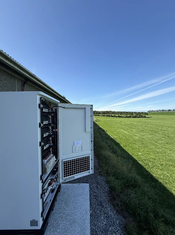 Batteriskap med åpen dør ved veggen på kyllinghuset, med grønne jorder og blå himmel i bakgrunnen