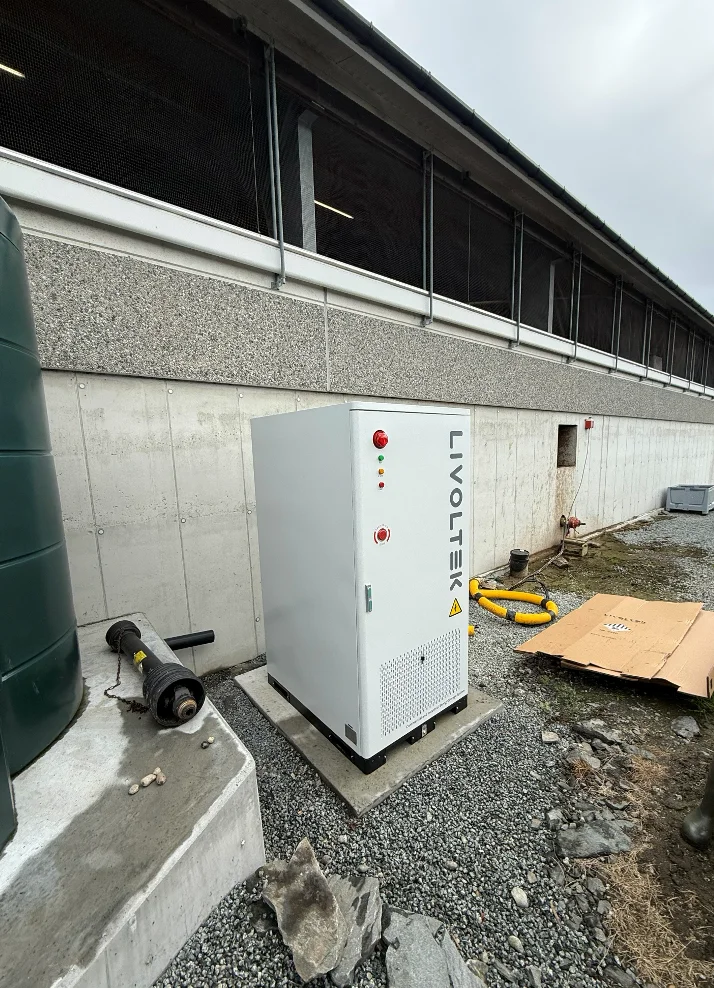Hvitt batteriskap fra Livoltek på et fundament ved den lange fjøsveggen på melkegården
