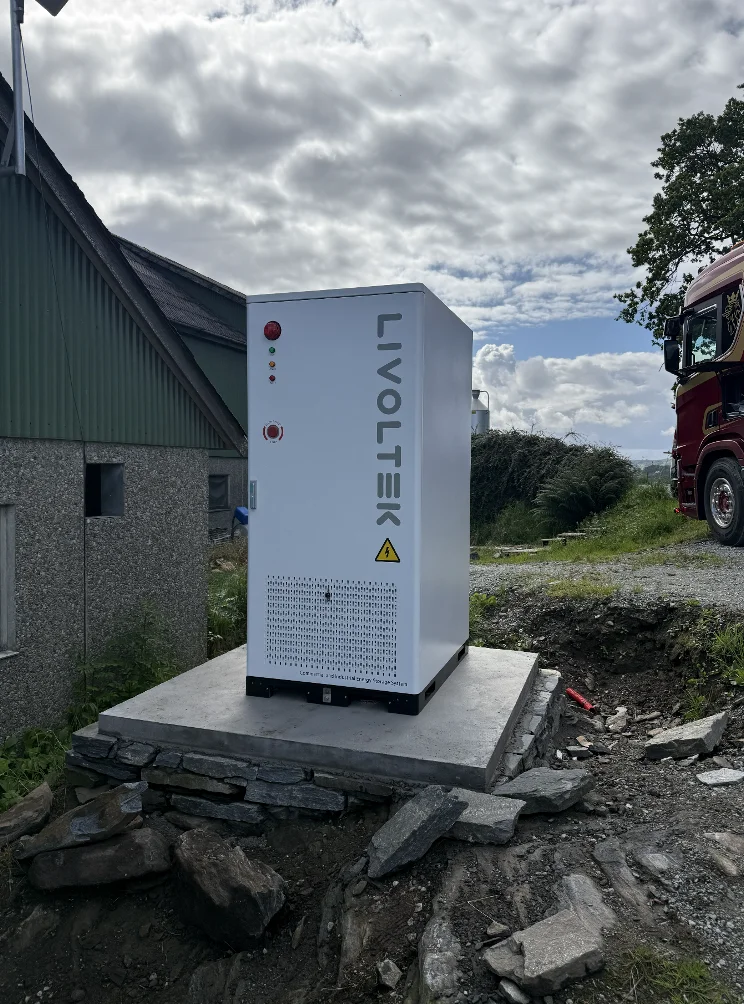 Hvitt batteriskap fra Livoltek på et fundament ved en grønn driftsbygning, med steinmur og gårdsvei rundt