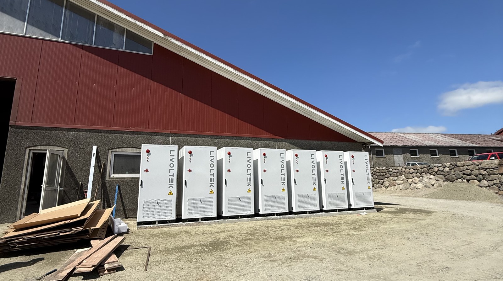 Sju hvite batteriskap montert på rekke langs veggen på en stor melkegård på Jæren, med blå himmel over