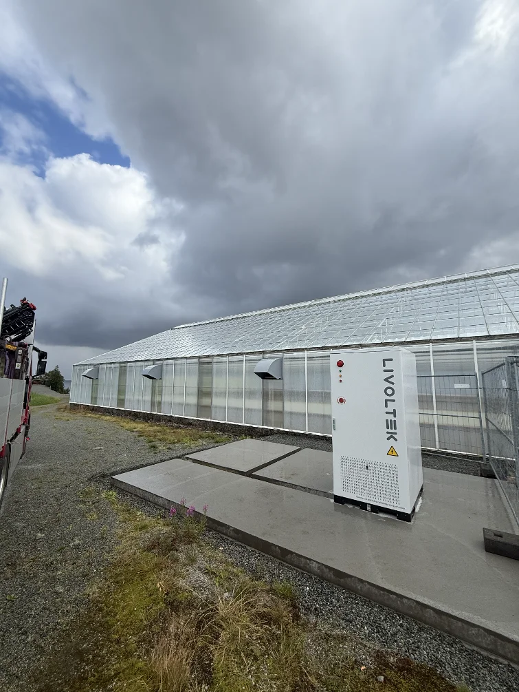 Hvitt batteriskap fra Livoltek på et betongfundament foran et veksthus, med overskyet himmel over